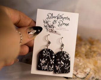 Può includere: Orecchini Blackthorn & Bone. Orecchini neri ad arco con disegni floreali e di uccelli argentati. Gli orecchini sono esposti su una carta bianca con il nome e il logo del marchio. Gioielli e decorazioni fatti a mano.