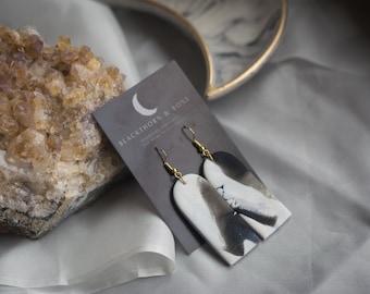 May include: A pair of black and white ceramic earrings with gold hooks. The earrings are shaped like a teardrop and have a marbled design. The earrings are displayed on a black card with the text "BLACKTHORN & BONE" in white.