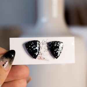 Op de afbeelding: Zwarte, schildvormige oorbellen met zilveren glitteraccenten. De oorbellen worden gepresenteerd op een witte kaart. Deze oorbellen maken deel uit van een sieradencollectie.