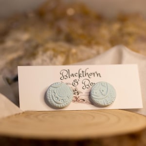 Könnte beinhalten: Hellblaue Ohrstecker mit aufwendigen geprägten Designs, präsentiert auf einer weißen Karte mit dem Text "Blackthorn & Bow". Die Ohrringe liegen auf einer Holzscheibe, mit unscharfem Hintergrund.