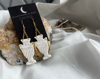 May include: A pair of white ceramic earrings shaped like urns with gold chain accents. The earrings are hanging from gold hooks. The earrings are on a black card with the text "BLACKTHORN & BONE HANDMADE JEWELLERY AND HOME ACCESSORIES".