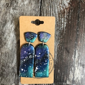 May include: A pair of galaxy-themed dangle earrings with a dark blue, green, and white colour scheme. The earrings are made of polymer clay and have a white splatter paint effect.