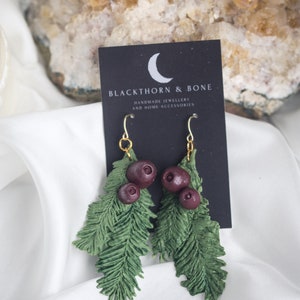 May include: A pair of earrings with green fir branches and dark red berries. The earrings have gold-coloured hooks and are displayed on a black card with the text "BLACKTHORN & BONE HANDMADE JEWELLERY AND HOME ACCESSORIES".