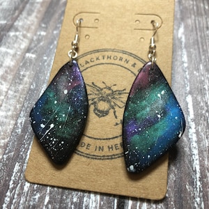 May include: A pair of galaxy-themed earrings with a black background and white, blue, and purple accents. The earrings are shaped like teardrops and have silver hooks.