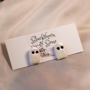 Può includere: Un paio di orecchini a forma di fantasma glitterati, con occhi neri e finitura iridescente. Sono presentati su un cartoncino bianco con la scritta "Blackthorn & Bone" e un'illustrazione di teschio.