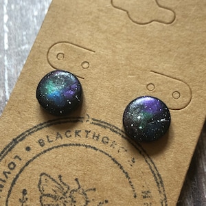 May include: A pair of galaxy-themed stud earrings with a black background and iridescent purple, blue, and green accents. The earrings are displayed on a brown card with a bee logo and the text "Blackthorn".
