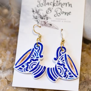 May include: A pair of white and blue ceramic earrings with gold-coloured hooks. The earrings feature a distinctive design with blue lines and patterns, and a touch of gold. They are displayed on a white card with the text "Blackthorn & Bone".