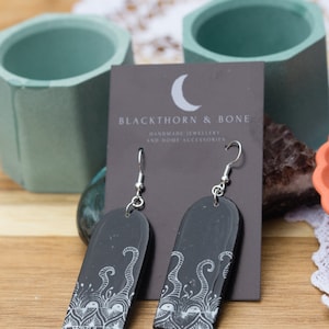 Könnte beinhalten: Schwarze und weiße Hängeohrringe mit schwarzem Hintergrund und weißen, wirbelnden Designs. Die Ohrringe befinden sich auf einer Karte mit der Aufschrift "Blackthorn & Bone Handmade Jewellery and Home Accessories".