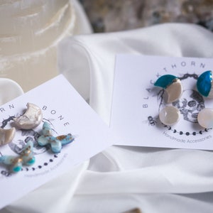 May include: Two sets of handmade earrings featuring crescent moon and butterfly designs. The earrings are made of white and blue resin with gold accents. The cards they are displayed on read "Blackthorn & Bone" and "Handmade Accessories".