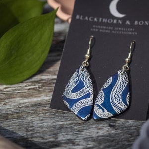 Puede incluir: Un par de pendientes de serpiente azul y blanco con ganchos plateados. Los pendientes se muestran en una tarjeta negra con el texto "Blackthorn & Bone Handmade Jewellery and Home Accessories".
