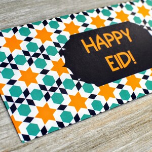 May include: A red and white envelope with a black and teal geometric pattern featuring orange stars. The envelope has a black label with the text "HAPPY EID!" in yellow lettering.