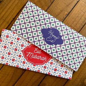 May include: Two decorative envelopes with a white background and a geometric pattern. One envelope has a blue and green pattern with the text "Happy Eid!" in a blue cloud. The other envelope has a red and blue pattern with the text "Eid Mubarak" in a red banner.