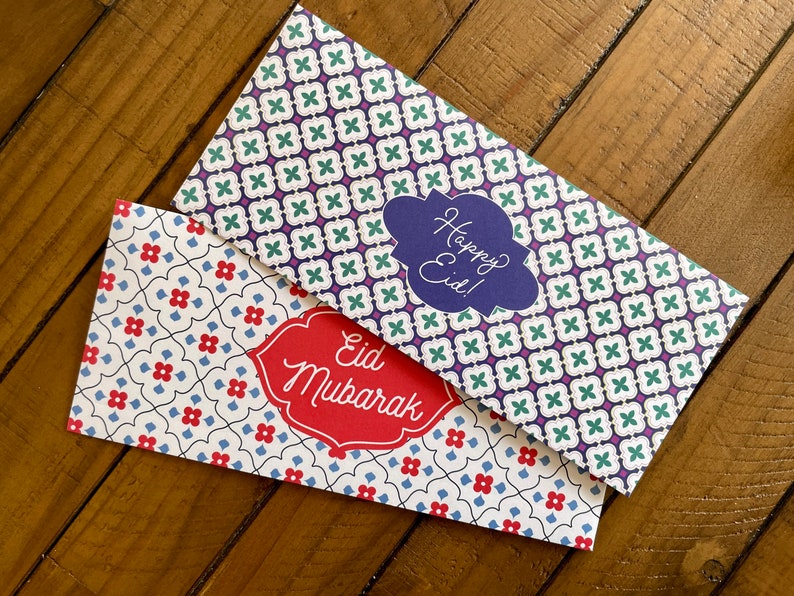 May include: Two decorative envelopes with a white background and a geometric pattern. One envelope has a blue and green pattern with the text "Happy Eid!" in a blue cloud. The other envelope has a red and blue pattern with the text "Eid Mubarak" in a red banner.