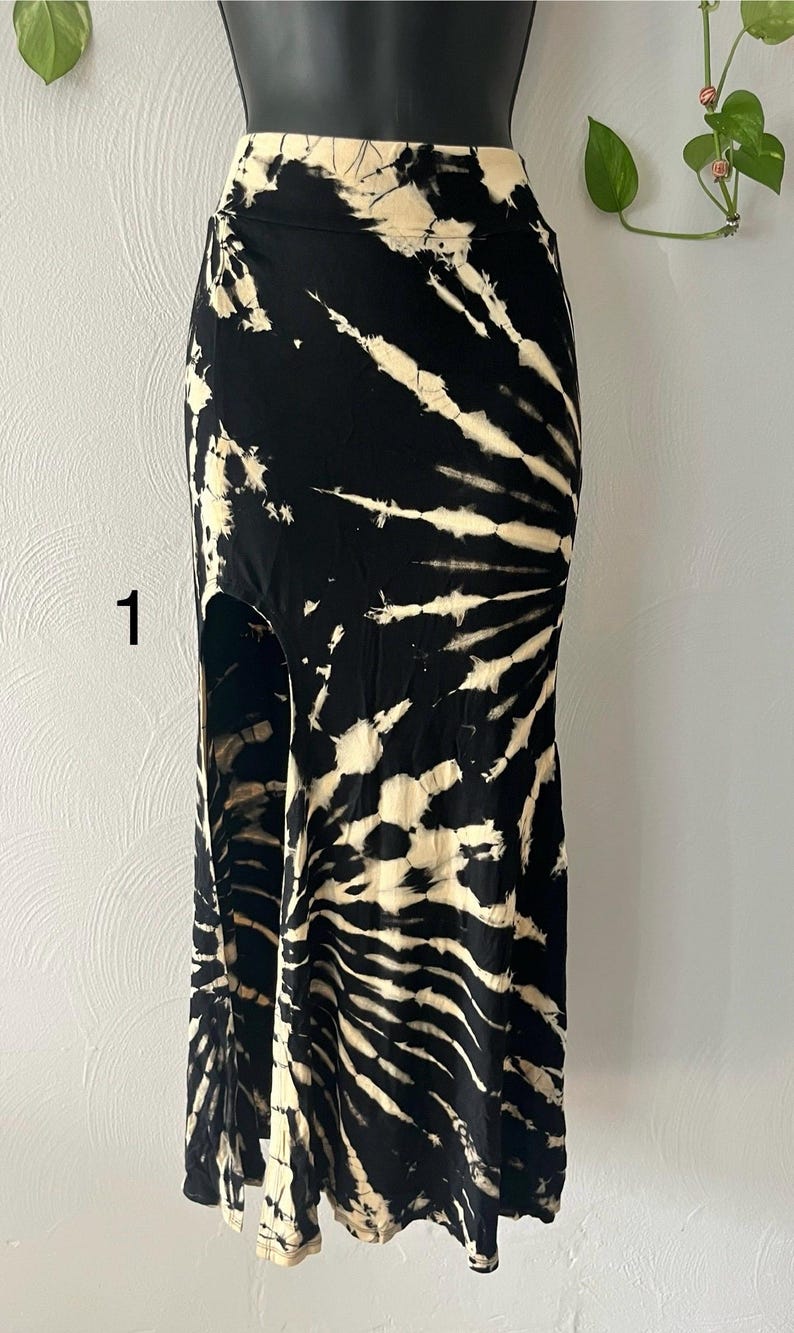 K&ouml;nnte beinhalten: Ein schwarzer und cremefarbener Maxirock mit Batikmuster. Der Rock hat eine hohe Taille und einen Seitenschlitz. Der Stoff hat ein wirbelndes Muster aus Schwarz und Creme, das einen einzigartigen Look erzeugt. Der Rock wird auf einer Schaufensterpuppe pr&auml;sentiert.