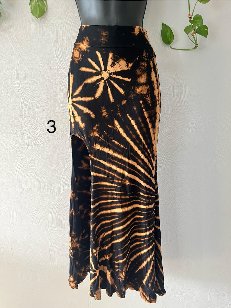 K&ouml;nnte beinhalten: Ein schwarz-beiger Batikrock mit hoher Taille und Seitenschlitz. Der Rock hat ein Sonnenmuster und die Zahl "3" ist auf der Vorderseite sichtbar. Der Rock wird auf einer Schaufensterpuppe pr&auml;sentiert.