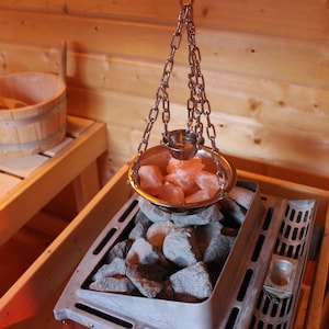 May include: A silver bowl filled with pink Himalayan salt crystals hangs from a chain over a metal sauna heater filled with gray rocks.