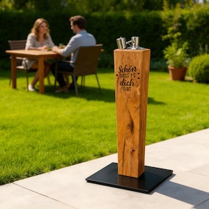 Könnte beinhalten: Eine Holz-Außenwasserfontäne mit Metallaufsatz und schwarzem Sockel. Auf der Fontäne steht der Text "Schön dass es dich gibt". Im Hintergrund sitzt ein Paar an einem Tisch.
