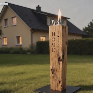 May include: A tall, rectangular wooden post with a natural grain and knots, featuring the word HOME and the German text die Liebe ist engraved on the front. A metal oil lamp burner with a lit flame is mounted on top, set on a black base.