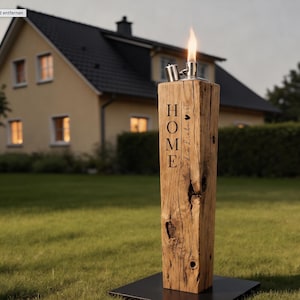 Pu&ograve; includere: Una torcia da esterno in legno rustico con una parte superiore in metallo e una fiamma. La parola "HOME" &egrave; stampata verticalmente sul davanti, con la frase "die Liebe ist" e un simbolo a forma di cuore. La torcia &egrave; posta su una base di metallo nero.