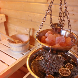 Może przedstawiać: Dwupoziomowa metalowa miska wisząca na łańcuchu w drewnianej saunie. Górna miska jest wypełniona różowymi kamieniami soli himalajskiej, a dolna miska zawiera wodę i suszoną lawendę.