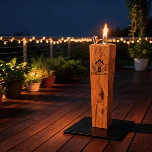 Könnte beinhalten: Eine Holzfackel für den Außenbereich mit Flamme, auf einem schwarzen Sockel. Die Fackel trägt den Text "LIEBLINGSORT" und ein Hauslogo. Der Hintergrund zeigt eine Terrasse, Pflanzen und Lichterketten, die eine warme Atmosphäre schaffen.
