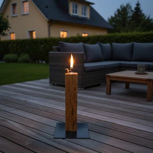 以下が含まれることがあります： 木製の柱と金属製のベースが付いた、点灯した屋外用オイルランプ。炎が明るく燃えています。ランプは木製のデッキに置かれ、背景には家と屋外用家具があります。