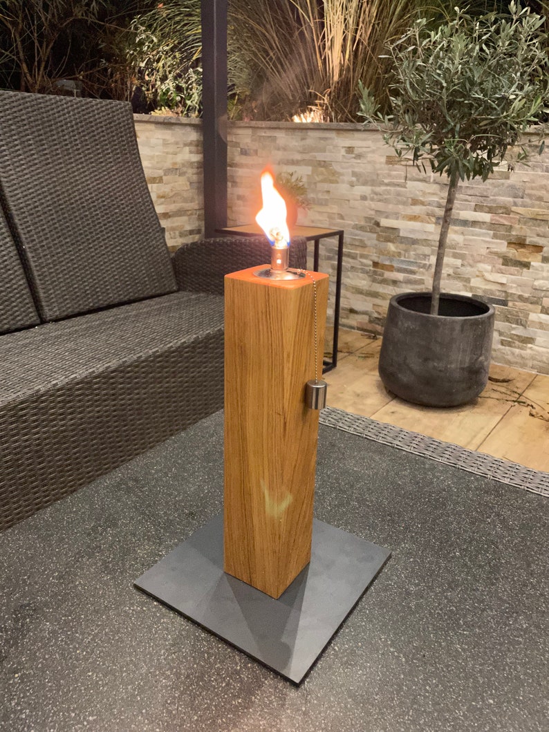 以下が含まれることがあります： 木製のベースと正方形の金属ベースを備えた、点灯した屋外テーブルトップの火鉢。炎はオレンジと黄色です。火鉢は灰色のテーブルの上にあります。暗い籐の椅子と小さな木が背景にあります。
