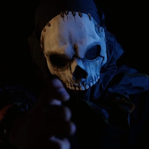 Może przedstawiać: Maska czaszki z zszytym wierzchem i ciemnymi oczodołami jest noszona z czarnym kapturem. Maska jest w kolorze złamanej bieli z ciemnym cieniowaniem wokół oczu i ust. Obraz jest ciemny z wyciągniętą ręką.