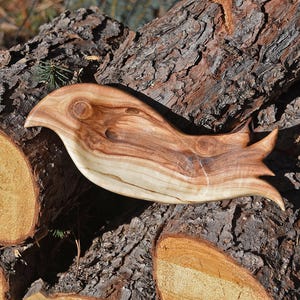 May include: A handcrafted wooden bird-shaped serving board. The bird is made of light and dark brown wood, with a smooth finish. The board is approximately 12 inches long and rests on a textured, dark brown log.