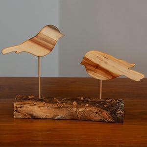 Può includere: Due sculture di uccelli in legno. Un uccello è in posizione eretta, l'altro è inclinato verso il basso. Entrambi sono marrone chiaro con venature più scure. Sono montati su sottili aste di metallo attaccate a una base di legno intagliato. La base è un pezzo di legno marrone scuro strutturato.