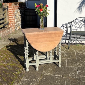 May include: A wooden drop-leaf table with a light brown tabletop and grey-painted legs. The table has a turned leg design. A clear glass vase with red and yellow flowers sits on the table. The table is approximately 76 cm high.