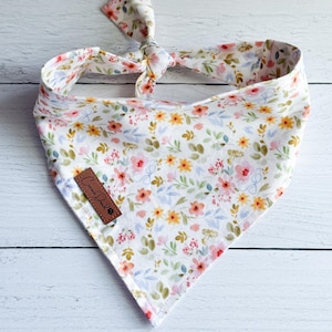 May include: A white dog bandana with a floral pattern in pink, yellow, and blue. The bandana is tied at the top and features a small brown leather tag with text. The fabric is soft and lightweight, perfect for pets.