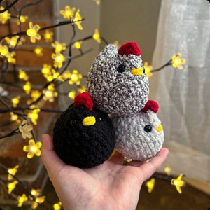 May include: Three crocheted chicks, one black, one white, and one gray, with red beaks and yellow eyes. The chicks are stacked on top of each other.