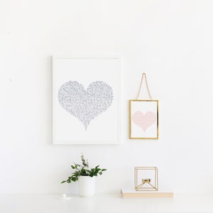 May include: Two framed prints of hearts, one with a black and white calligraphic design and the other with a pink heart design. The prints are hanging on a white wall with a small plant in a white pot on a white surface.