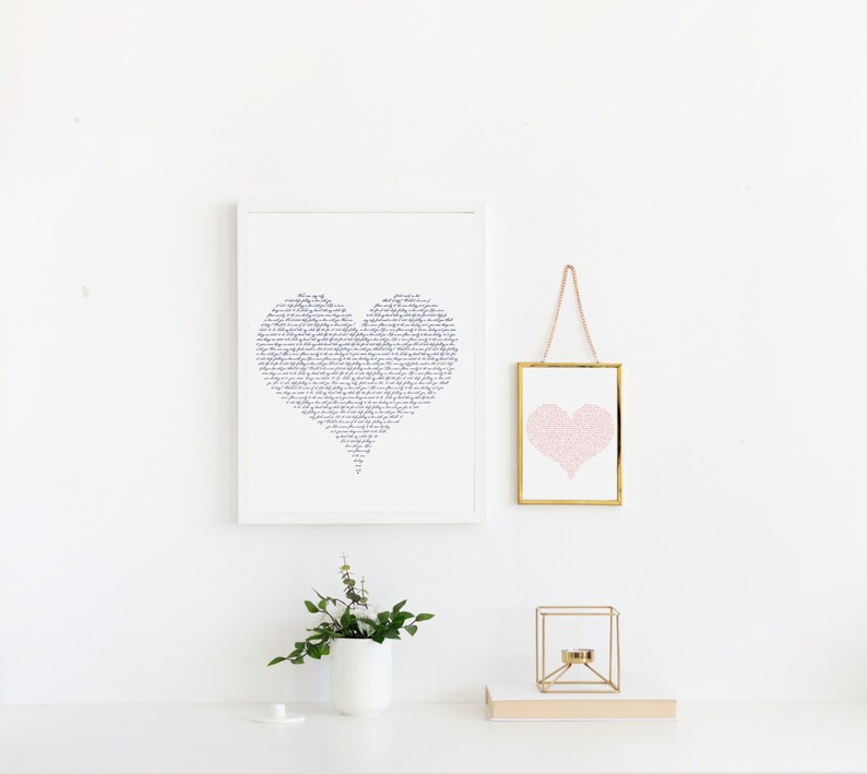 May include: Two framed prints of hearts, one with a black and white calligraphic design and the other with a pink heart design. The prints are hanging on a white wall with a small plant in a white pot on a white surface.