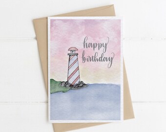 Op de afbeelding: Een aquarel-illustratie van een vuurtoren op een rotsachtige kust met een roze en blauwe lucht. De tekst "happy birthday" is in cursief op de kaart geschreven.