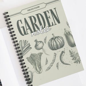 May include: A spiral-bound notebook with the words "GARDEN notes" on the cover, featuring illustrations of vegetables. The cover is a light green color. The notebook is open on a white surface, with a gold paper clip and sunglasses nearby.