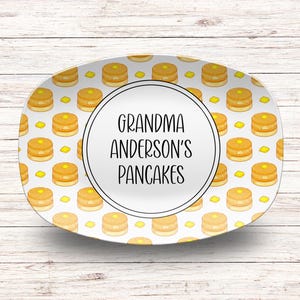 Puede incluir: Plato ovalado con un patrón de panqueques con mantequilla sobre un fondo blanco. El centro tiene un círculo blanco con el texto "GRANDMA ANDERSON'S PANCAKES" en negro.
