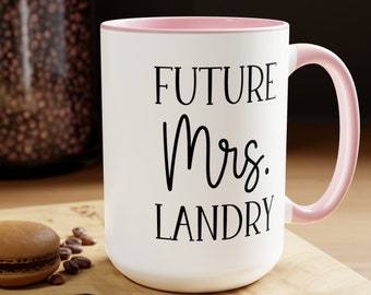 May include: White ceramic mug with a pink rim and handle. The mug has black text that reads "FUTURE Mrs. LANDRY".