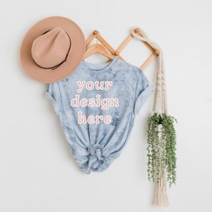 May include: A light blue tie-dye t-shirt with the text "your design here" in pink lettering. The shirt is hanging on a wooden rack with a brown hat and a macrame plant hanger.