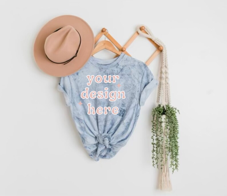 May include: A light blue tie-dye t-shirt with the text "your design here" in pink lettering. The shirt is hanging on a wooden rack with a brown hat and a macrame plant hanger.