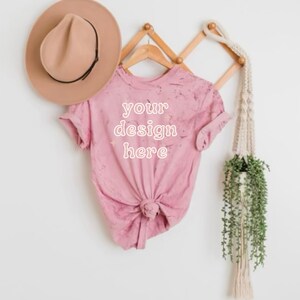 May include: A pink t-shirt with a distressed look, hanging on a wooden rack. The shirt has the text "your design here" printed on the front.