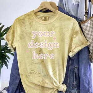 May include: A yellow tie-dye t-shirt with the text "your design here" in pink. The shirt is hanging on a wooden hanger.