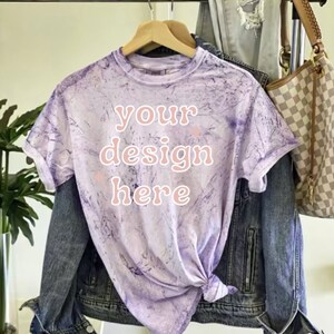 May include: A purple tie-dye t-shirt with the text "your design here" in pink. The shirt is hanging on a wooden hanger and is displayed with a denim jacket and a brown and white checkered handbag.