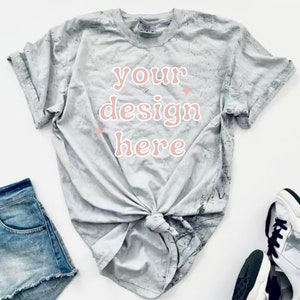 May include: A gray tie-dye t-shirt with the text "your design here" in pink. The shirt is knotted at the front and is laying on a white surface.