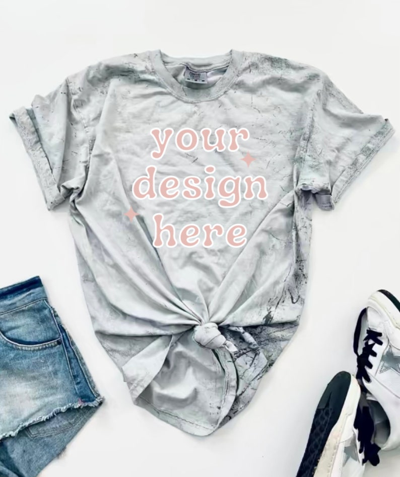 May include: A gray tie-dye t-shirt with the text "your design here" in pink. The shirt is knotted at the front and is laying on a white surface.