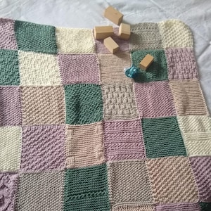 Könnte beinhalten: Eine gestrickte Patchwork-Decke mit Quadraten in Rosa-, Grün- und Cremetönen. Die Decke ist mit verschiedenen Strickmustern gearbeitet, wodurch ein strukturierter Look entsteht.