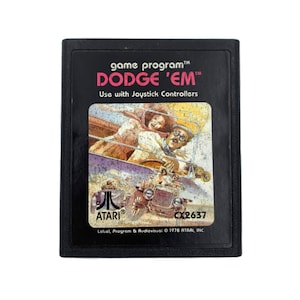 May include: A black Atari game cartridge with a colourful illustration of a car and people on the front. The text "game program™ DODGE 'EM™" is printed in red on the front of the cartridge. The text "Use with Joystick Controllers" is printed in black below the title. The text "ATARI® CX2637 Label, Program & Audiovisual © 1978 ATARI, INC." is printed in black on the bottom of the cartridge.