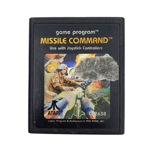 May include: A black Atari game cartridge with a colourful image of a spaceship and a pilot. The text on the cartridge reads "game program™ MISSILE COMMAND™ Use with Joystick Controllers ATARI® C2638 Label, Program & Audiovisual © 1981 ATARI, INC."