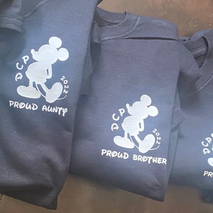 May include: Five dark grey t-shirts with a white silhouette of Mickey Mouse and the text "DCP 2022" and "Proud" followed by a family member title. The titles are "Alex", "Auntie", "Brother", "Dad", and "Grandma".